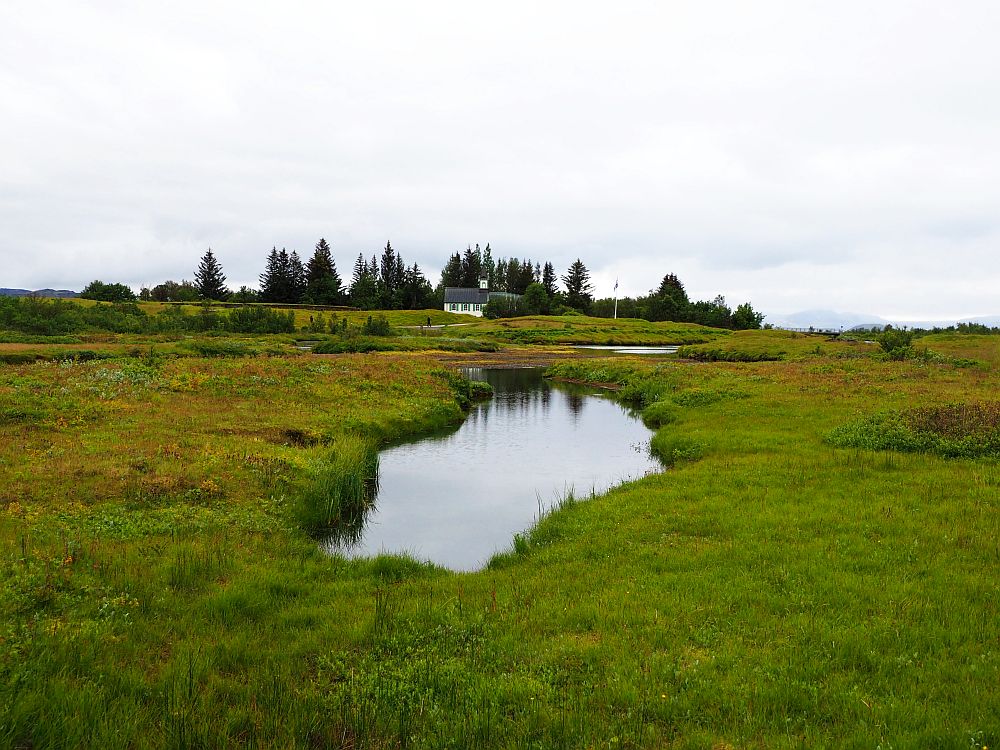 On the horizon, the white church is partly visible, with a row of trees behind it. Nearer by: green fields and a small pond in the center.