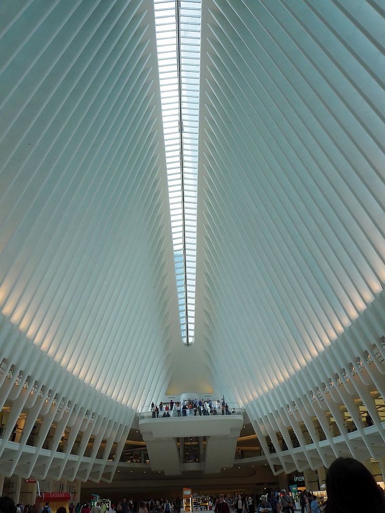 A soaringly high ceiling extends ahead, with a large stairway and some very small people visible at the far end. Along the top, the peak of the ceiling, is a double row of windows letting light in. The side walls are ribbed with vertical ribs.