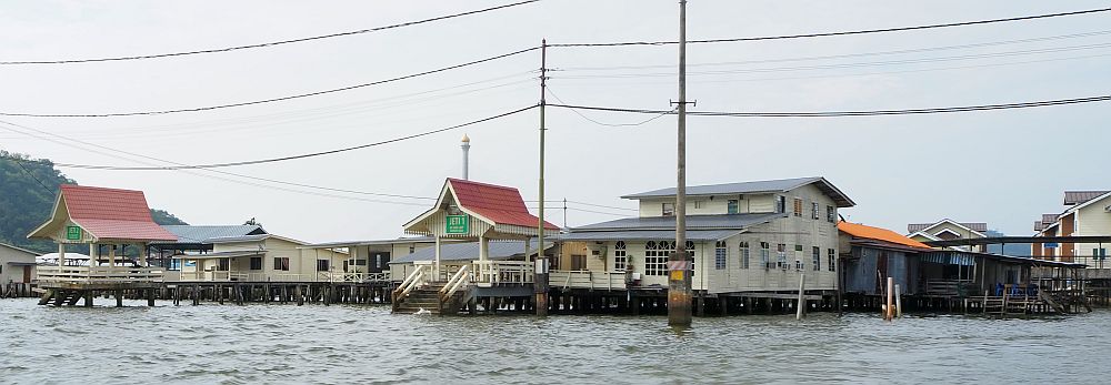 White houses on stilts over the water, and two small roofed quays extend out from them. Electric lines cross the top of the picture on poles in the water.