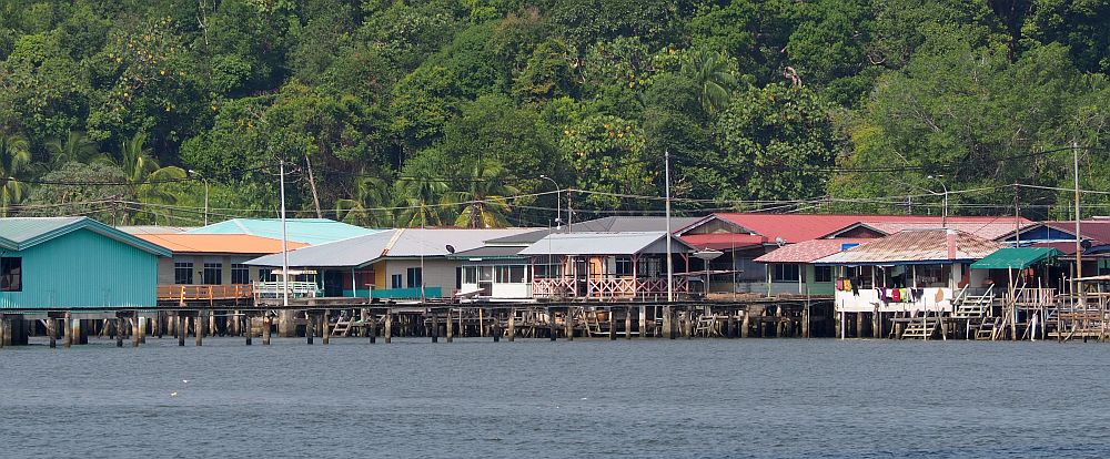 A cluster of houses on stilts over the water, some painted in bright colors, all of them just a single story with wide verandas. One has a lot of laundry hanging from the veranda. Behind is a tree-covered hill. 