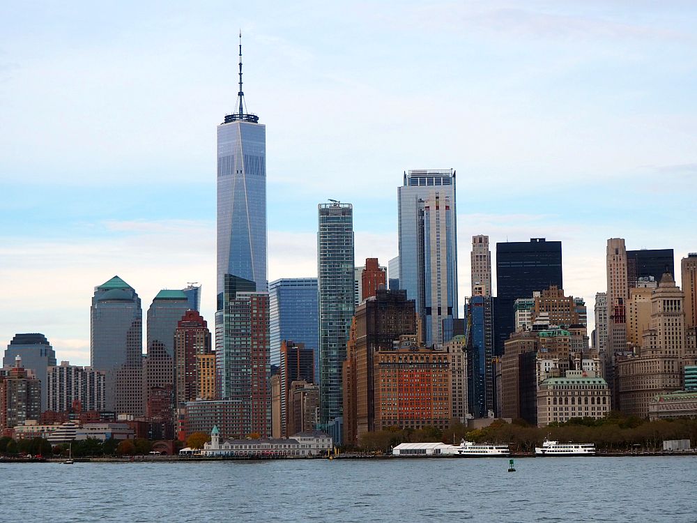 Across the water, a city skyline: the southern part of Manhattan. The tallest building is silver with glass and has a tall mast on the top: the One World Trade Center Building.