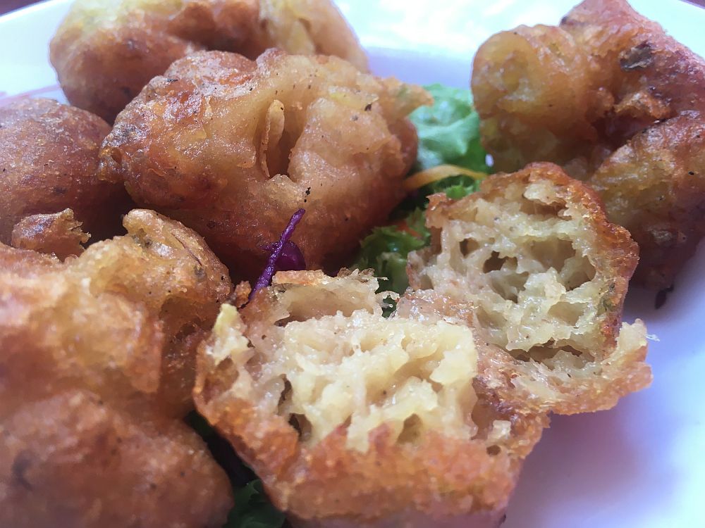 Close-up of a plate of fritters: small lumpy fried balls. Once is broken open to reveal a bread or cake-like consistency inside.