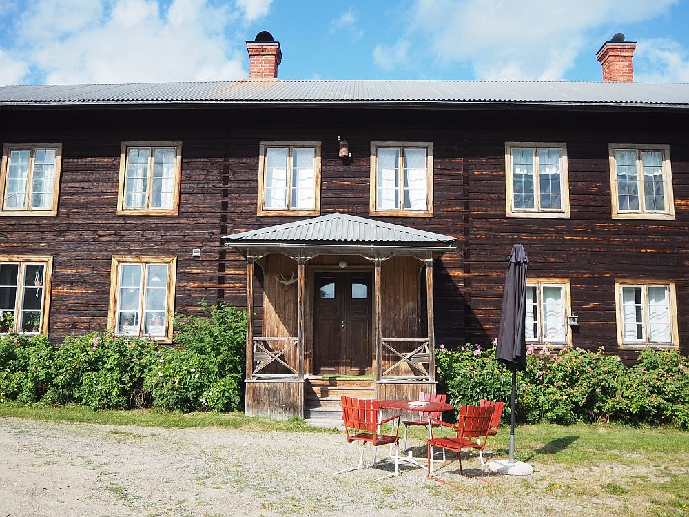 A brown wooden house with two stories and evenly spaced, almost square windows. The entrance is square and right in the middle.