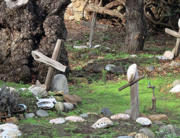 Two graves in the foreground, each with stones on it and a very crude wooden cross. The cross on the right has a large oyster shell hanging from it. There is a big old tree trunk behind the left-hand grave and another grave is visible in the background on the right.