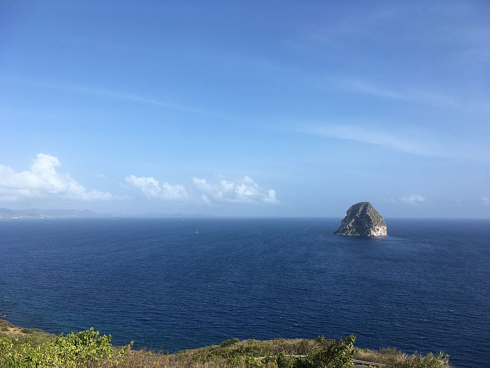 A view over  a calm blue ocean with one bare, pointed rock sticking up out of it.