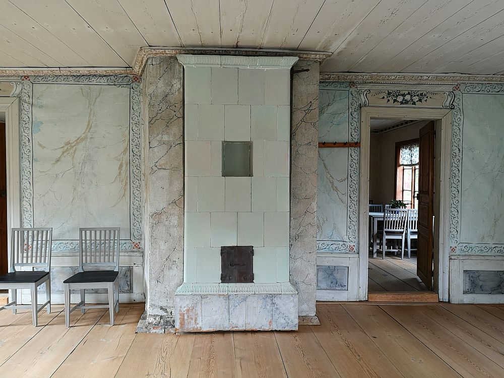 The walls here are light blue, with brown fake marbling across each. The edges have a simple pattern painted on them in a darker blue. A flowery garland in white and blue above the doorway. IN the center is a tiled furnace in a very simple floor-to-ceiling style.