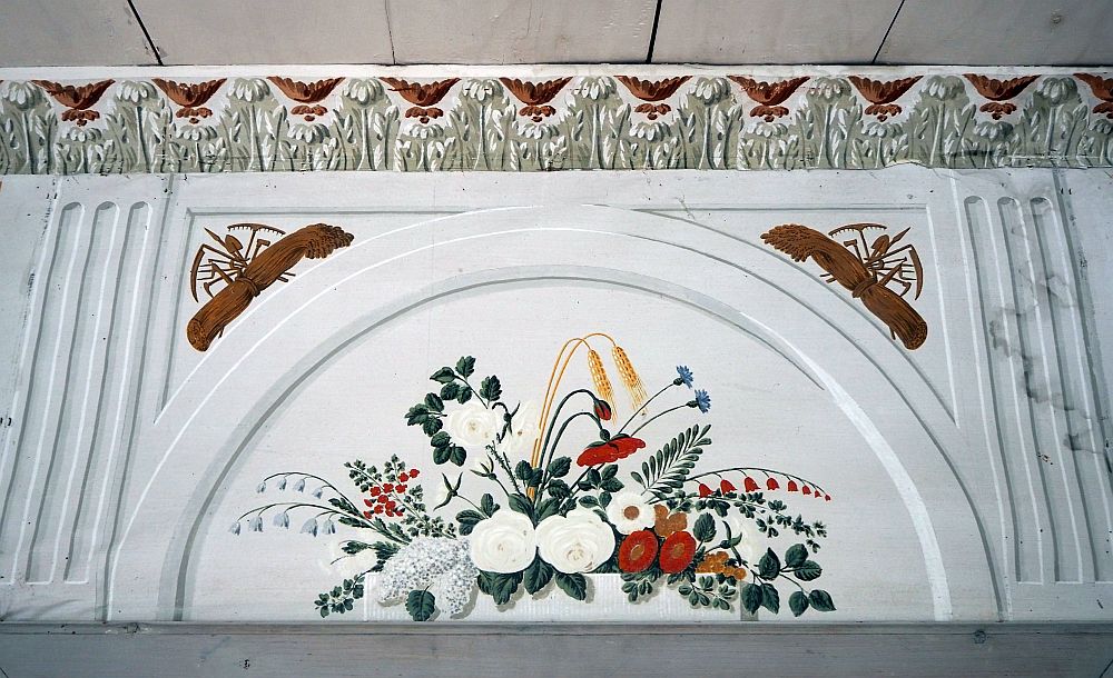 A half-circular arch over the door. White background with an arrangement of wildflowers in white, red and green painted on it. Top left and right are bundles of hay above the arch, each with a collection of agricultural implements. Above the arch is the molding along the edge of the ceiling, which is painted with a repeting pattern of garlands in brown and olive green.