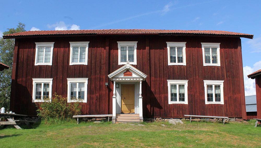 A simple red-painted house with white trim around the windows. The doorway is set right in the middle with a small roof over the entry. Two stories. Two windows on either side of the door and 5 windows upstairs.