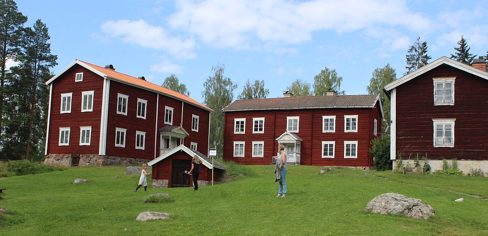 This photo shows all three red-painted houses with white trim, set in three sides of a square. In front is a little shed half in the ground.