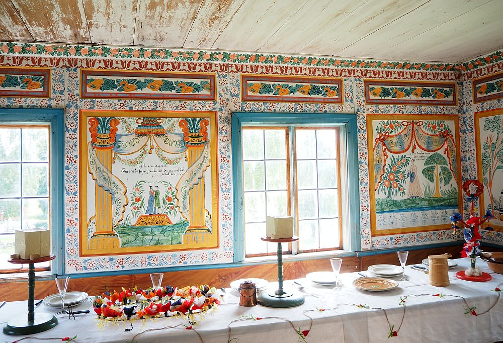 A long table at the bottom of the photo has a simple table setting with occasional colorful arrangements. Behind them are two windows with the walls between and above them completely filled with paintings: Large square murals between and next to the windows show scenes filled with flowers. The center one has a couple on a boat, I think, in the center and the other shows a woman standing among flowering trees. Around the murals and above them are ornate flowery patterns, and a different pattern follows the molding next to the ceiling. By contrast, the ceiling is plain white.