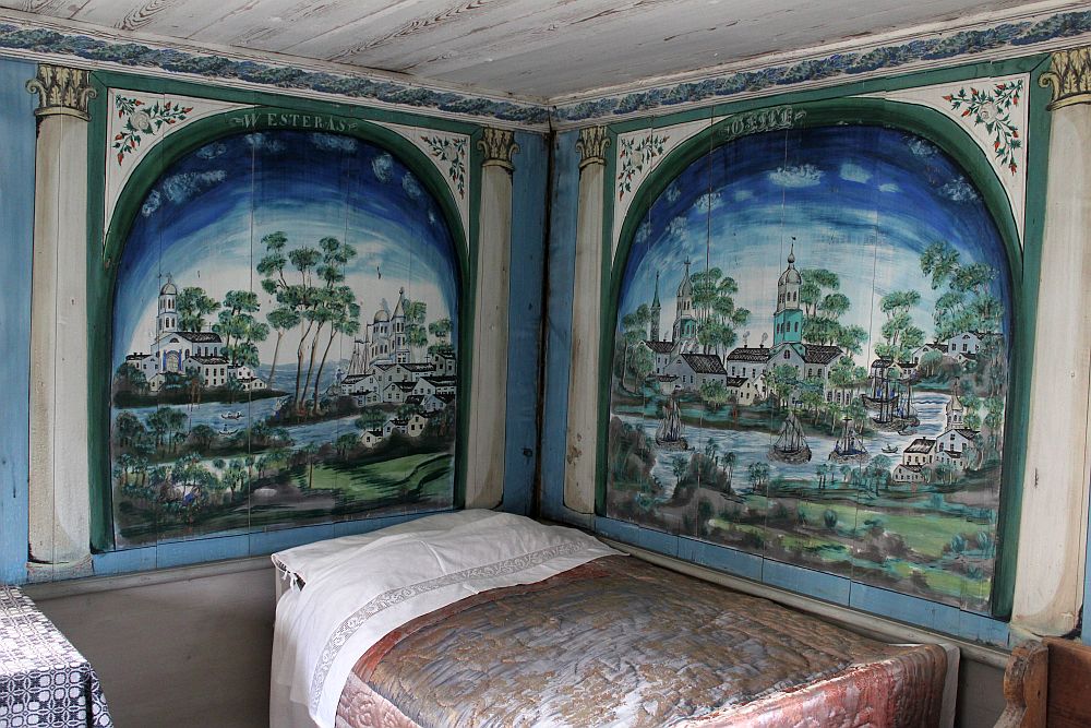 A bed in a corner. Above it, on each wall, a large painting filling each wall, arched at the top. Each painting shows a scene of white buildings, a church, a waterway with boats. The sky above is blue and the top edges are extra blue. 