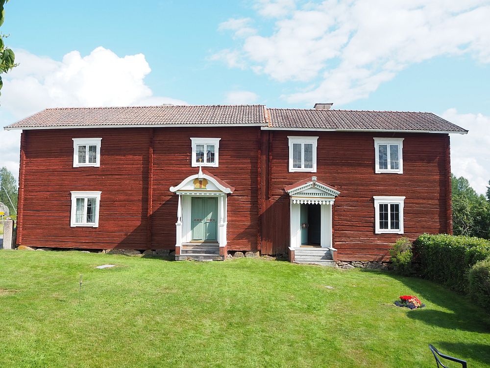 Each half has a doorway toward the center where the two houses meet, with twoo different small porticos. Each house has 3 windows: One next to the door and two on the upper story. The house is wooden and painted dark red.