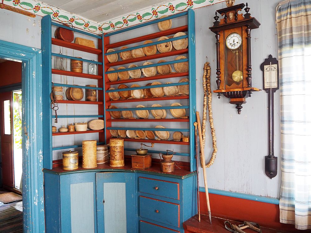 In the corner is a cupboard with cabinets and drawers below and plate rails all the way to the molding above. The whole thing is painted in bright blue, light blue and deep red. Next to it a large clock hangs on the wall. A flowery molding lines the ceiling above.