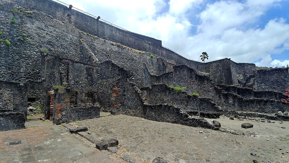 A high wall with crumbling walls sticking out at right angles to it, showing where the rooms of this building used to be. All seem to be made of stone.