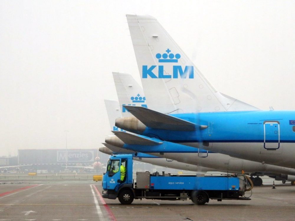 A row of KLM planes parked on the runway: Just their tails, in a neat row, are visible here, with one service truck parked under the nearest one.
