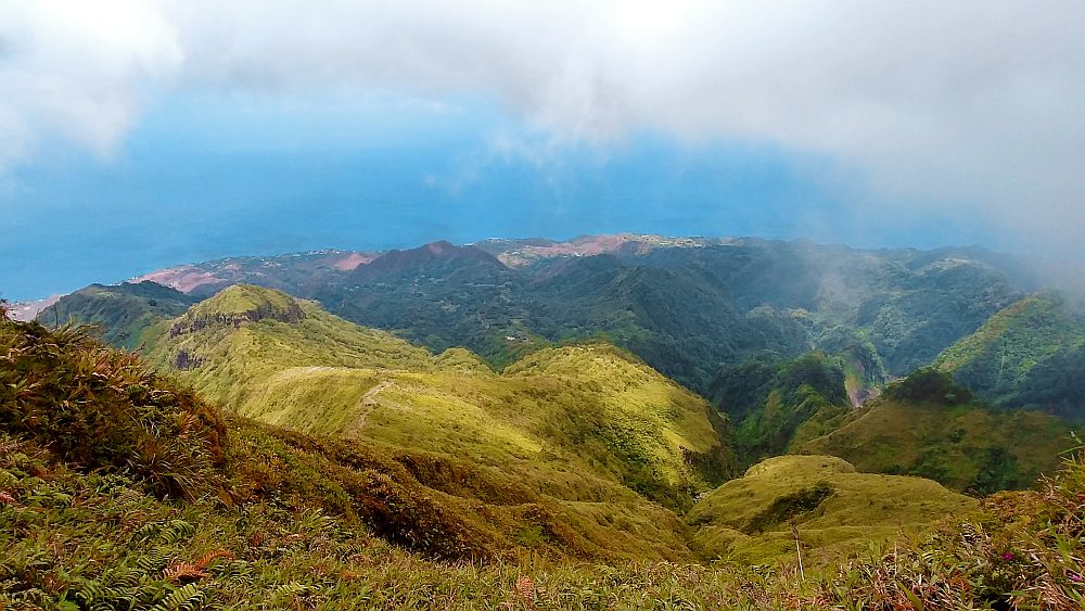 the view shows green hills nearby, covered with some sort of low growth. IN the distance, more hills covered with darker green growth, and beyond that, through a bit of thin cloud, the sea is visible.