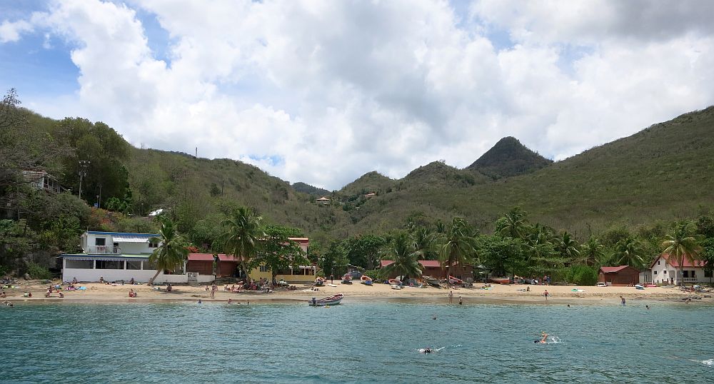 A view of a small beach as seen fro m the water, hills covered in greenery rise behind it. The beach has a lot of palm trees along it and only a few small buildings. A few people swim in the water or stand or lie on the beach.