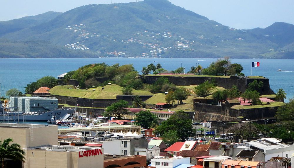 A view across an area packed with low-rise buildings to a higher point of land with water beyond it. The point has walls around and on top of it and a French flag flying from its highest point. On the other side of the water is another shore with a mountain.