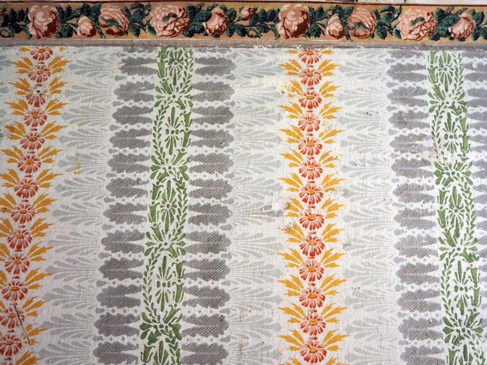 The stenciled patters repeat vertically right to the ceiling molding, which is painted in pink roses with green leaves. The patterns also repeat horizontally: left to right: a vertical line of red flowers with yellow leaves, then a light grey spray of leaves, a dark grey spray of leaves, a green pattern of flowers and leaves, then back to dark gray, light gray and then the red and yellow again.