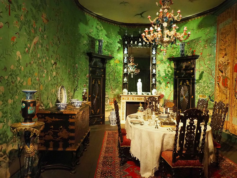 The walls are predominantly green with flowery images painted onto them. The table is elegantly set, with a white tablecloth and all the usuall table settings. The chandelier is rather chunky-with each candle in an ornate glass holder. Against the wall on the left is a large piece of furniture holding several pieces of Chinese porcelein.