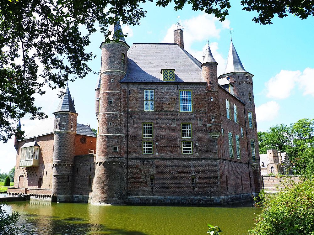 The green water of the moat in the foreground. Behind, the castle, red brick, about 4 stories high, with turrets on each corner. 