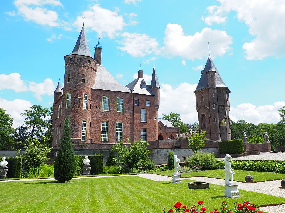 IN the foreground, very tidy green grass lawn with statues here and there. Beyond, the castle: red bricks, with a large tower in the near corner and smaller little turrets here and there, as well as a separate tower on the right. 