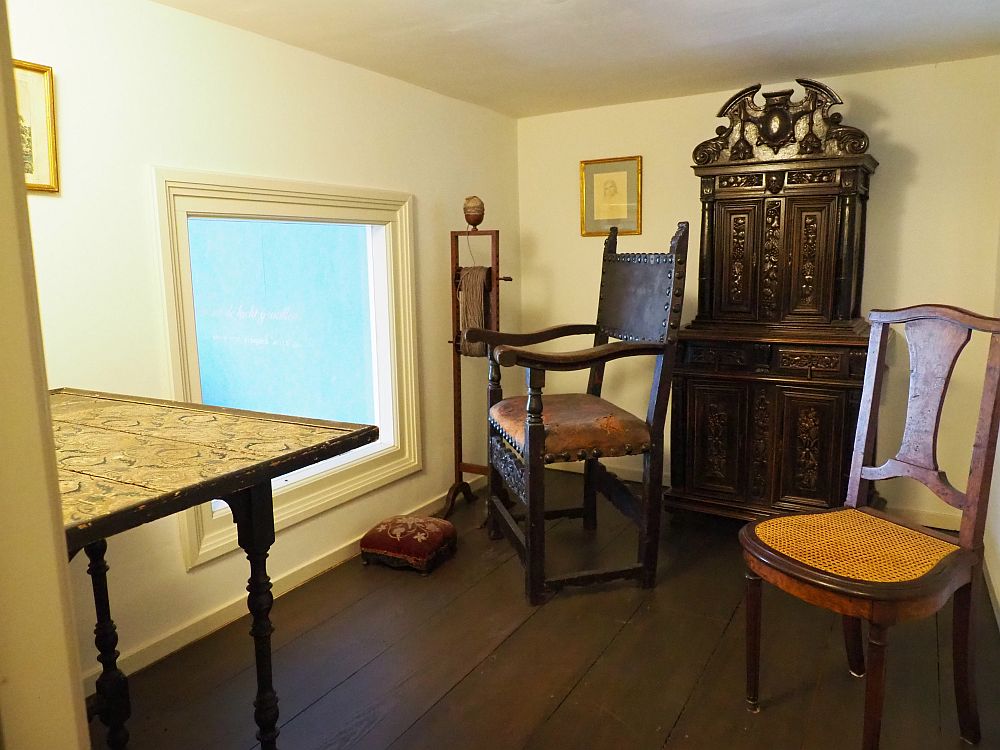 A low-ceilinged room, quite plain. The window is low near the floor on the left. Plain wood flor with two simple wooden chairs, a small carved secretary and a table. 