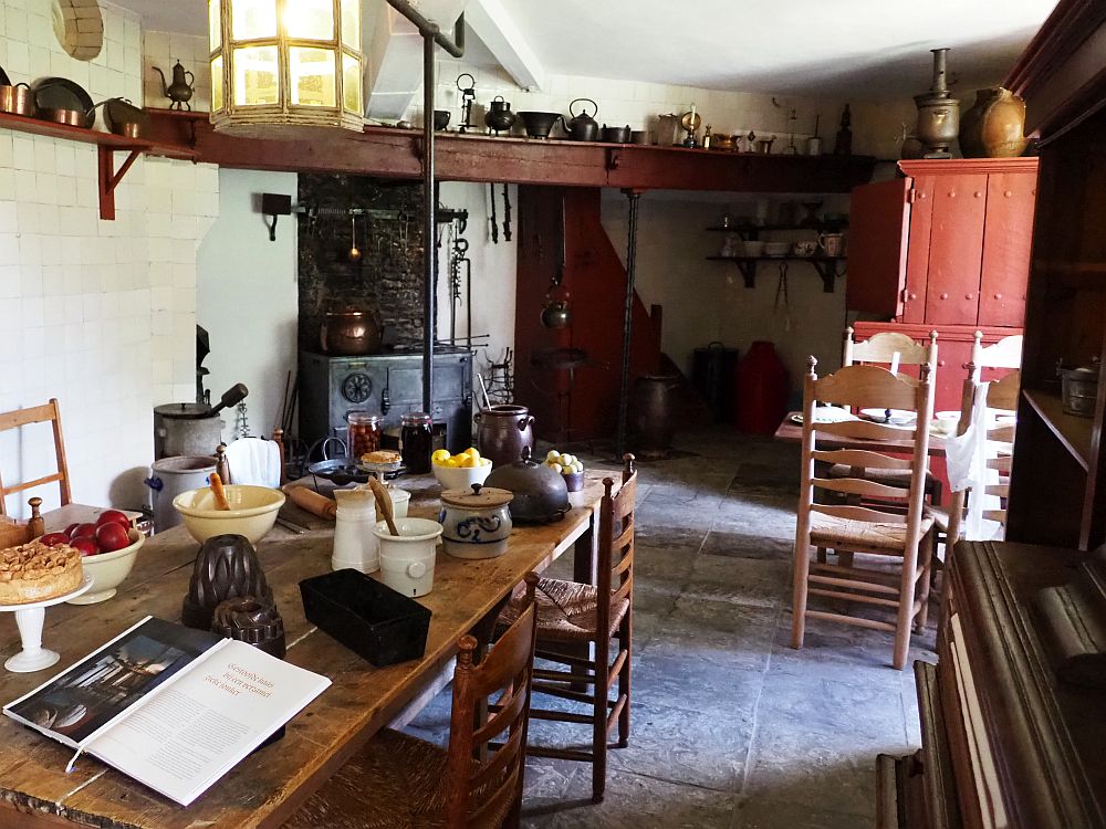 Foreground, a long wooden table with various food preparation tools on it like morters and pestles. Beyond, a cast-iron stove in the far corner. On the wood beam above it are various other tools: kettles and pots and such. On the right halfway down the room part of another wooden table with chairs around it is visible, and a large cupboard beyond that. The floor is rough stone and the walls are plain white.