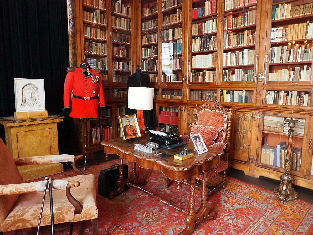 A large wooden desk with an upholstered seat behind it and another in front of it. A typewriter and some framed pictures on the desk and a bright red uniform on a stand next to it. Behind the desk the wall is filled with glass-fronted wooden shelves filled with books.