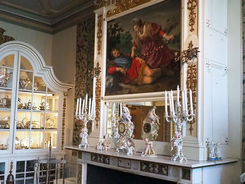 On the right is a grand mantel with two clocks in the center made of white porcelein, very flowery. One other end is a white porcelein candelabra with multiple candles. Behind the clocks is a mirror and above that a painting with two figures, one liying down, another leaning over the first on her knees. Around the painting is gold-painted edging. Left of the mantal is a large glass-fronted cupboard with a rounded top, lit from inside, where various china and silver is on display.