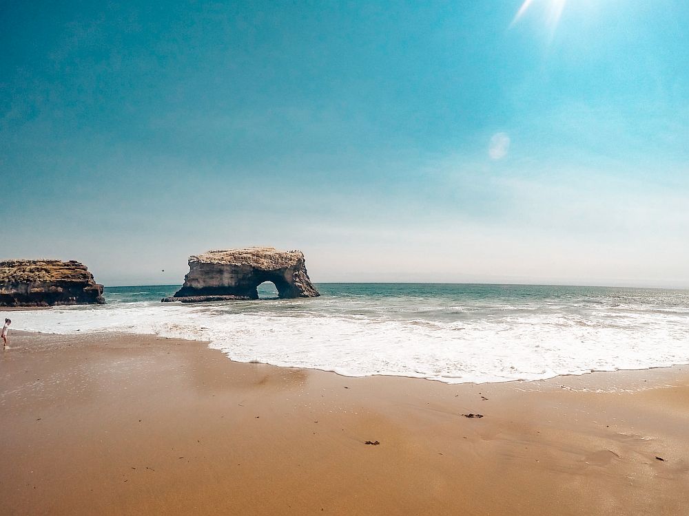 A view from a beach onto the sea, white foam from a wave along the sea's edge. Not far out to sea is a chunk of rock shaped like an arch.