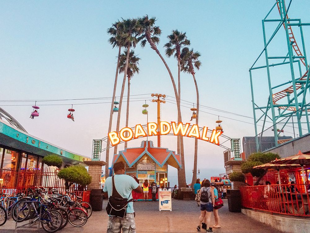 Looking down the boardwalk: a small kiosk straight ahead with an arched neon sign above it that reads "boardwalk". TO the right a small part of a roller coaster track is visible. Behind the kiosk in the center is a group of about 6 very tall narrow palm trees. Beyond that is a horizontal cable where seats dangle along its length.