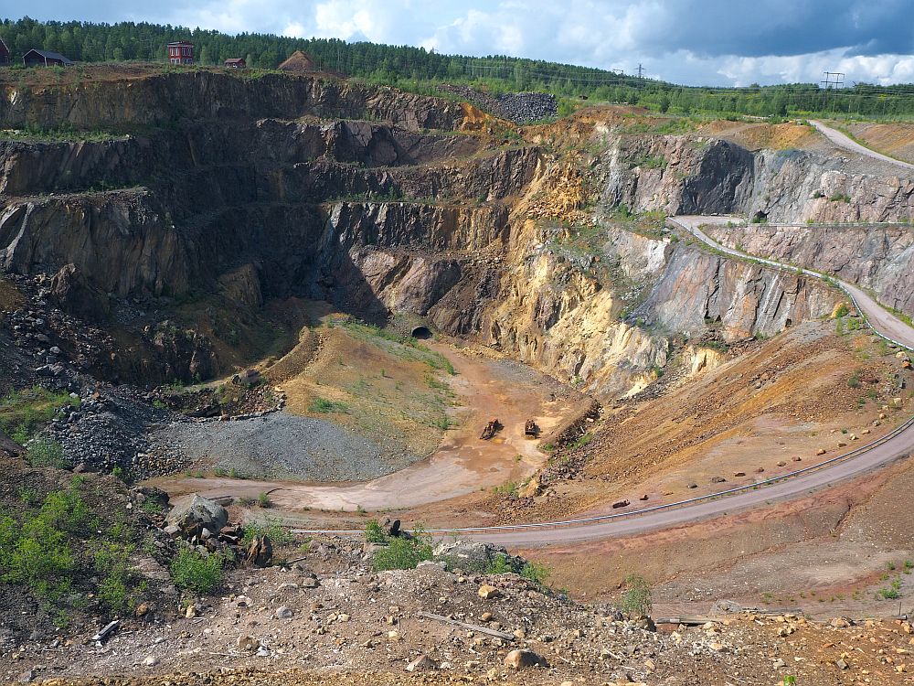 A deep pit at Falun Copper Mine, showing bare rock around the sides, some of which is reddish. A dirt road snakes down to the bottom and two tractors are visible, very tiny, at the bottom.