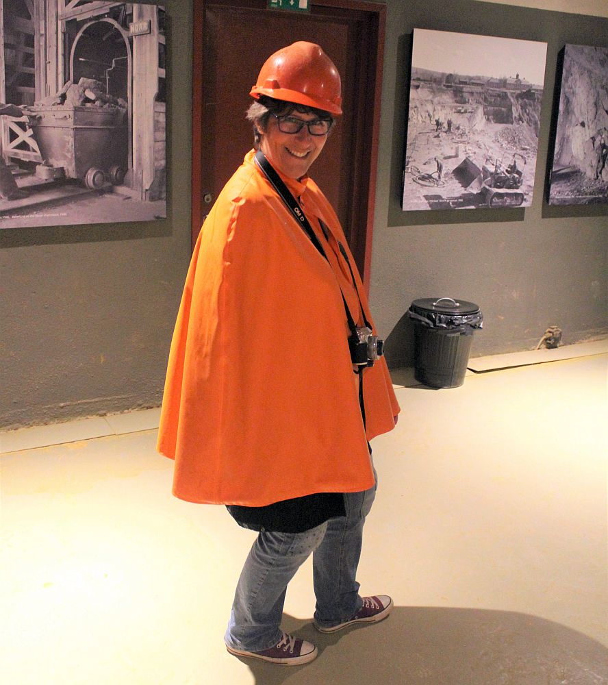 A photo of me, smiling at the camera. I wear an orange hardhat and an orange rain poncho, jeans and sneakers. A camera hangs around my neck.