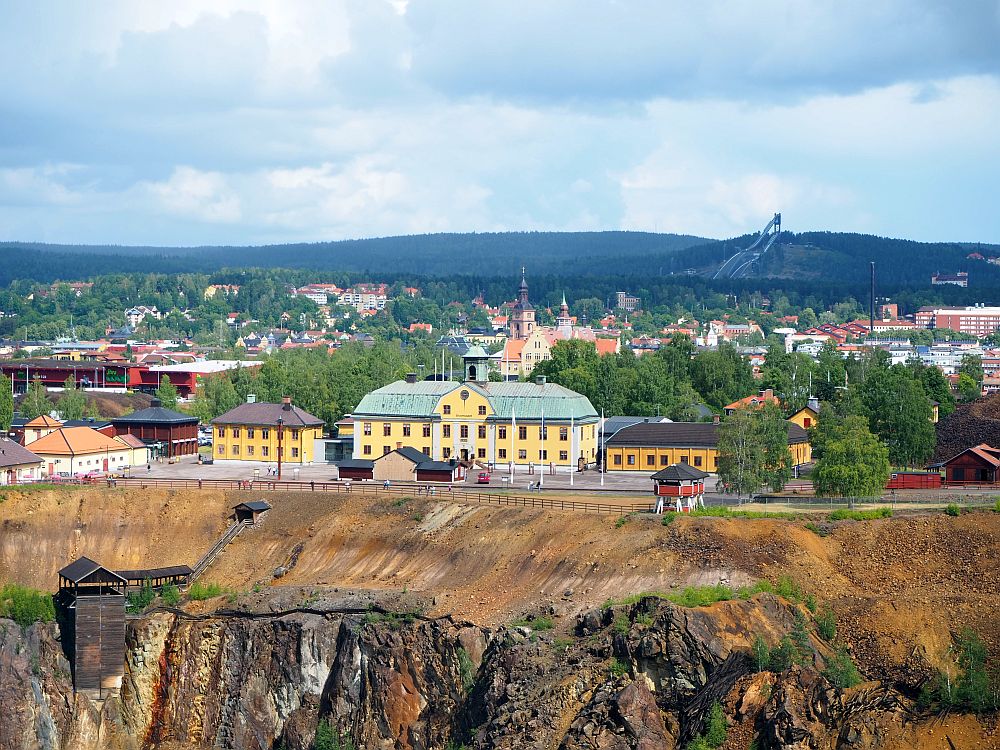 The side of the pit is visible in front: a rock wall. A small brown roofed structure hangs on the edge of it, with a stairway down to it from the top edge of the pit. Beyond that is the mine office building, painted yellow with a green roof. Smaller yellow buildings on either side of it: 2 stories on the left, single-story on the right. A church peeks from behind the mine building and a whole village can be glimpsed behind that. 