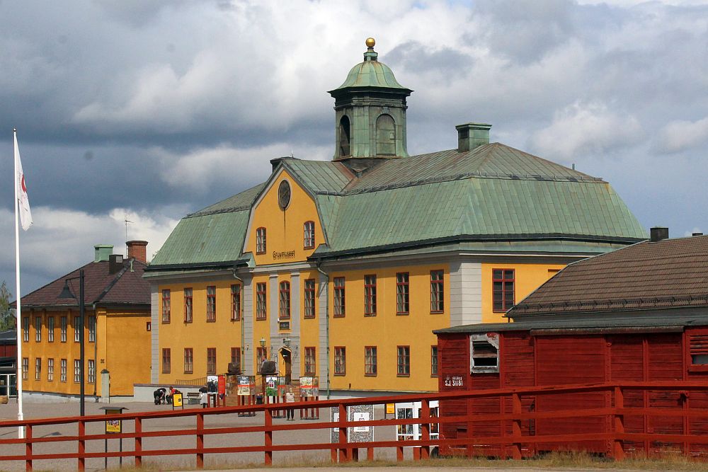 The mine building is a 3-story yellow-painted structure, symmetrical, with neat rows of windows on the ground floor and first floor. A door in the middle and 5 window on each side. The top floor is under a green roof, and only two windows near the center show, with a round window centered above them.