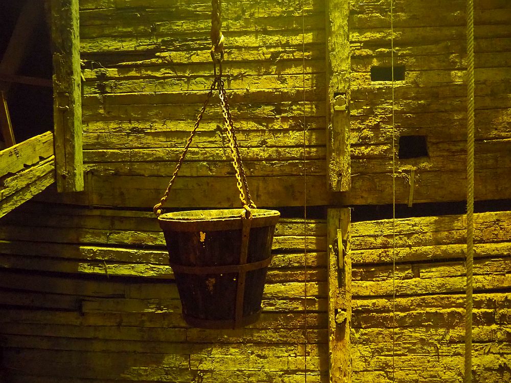 A pail hangs on a chain that connects with the pail at 3 points. Behind the pail, the wall is made of parallel horizontal strips of wood with much larger wood beams going horizontally. The whole scene has a greenish cast with indirect light from above.