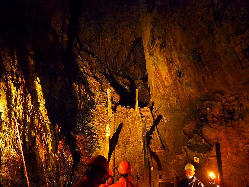 The heads of several people are visible at the bottom of the photo, wearing hardhats of orange or yellow (the tour guide). Indirect lights reveal two parallel stairways up a slope to a tunnel that disappears into darkness. The stairways are wooden with a broken railing on the side between the two stairways. ON the outside is rock walls. The stairway on the left is partly covered in rubble The one on the right has a broken tread toward the top and rubble toward the bottom.