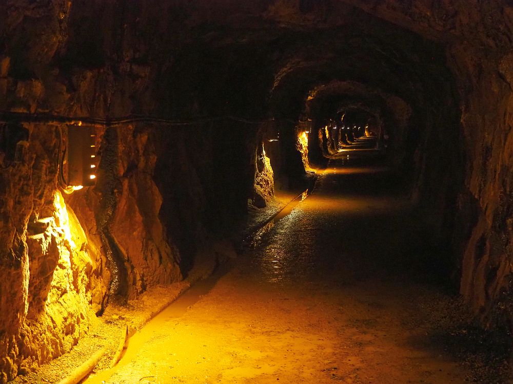 Looking down a long dark tunnel, with a downward-shining light perhaps every 5 meters going off into the distance. The nearest lamp shows that the wall is rough cut stone and that the floor is fine gravel and quite wet.