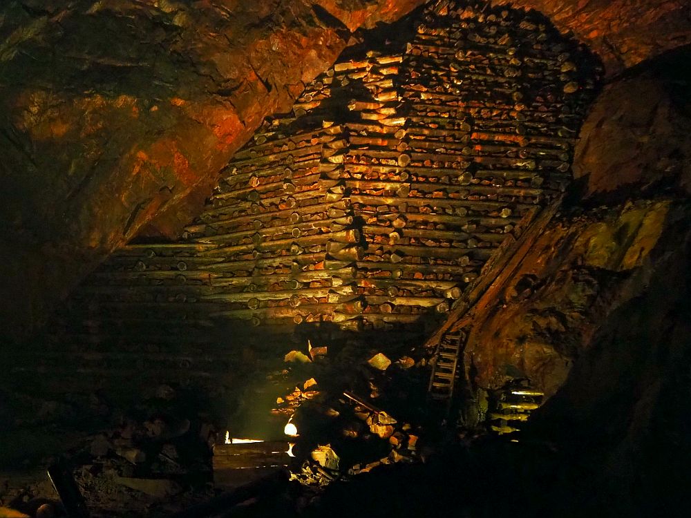 Inside a large cave, stone walls on left and right. The far wall, though, is large round logs laid horizontally, like a log cabin, but with spaces between the logs, which show rocks. All of it is a bit reddish. At the bottom is an old wooden ladder and lots of rocks.