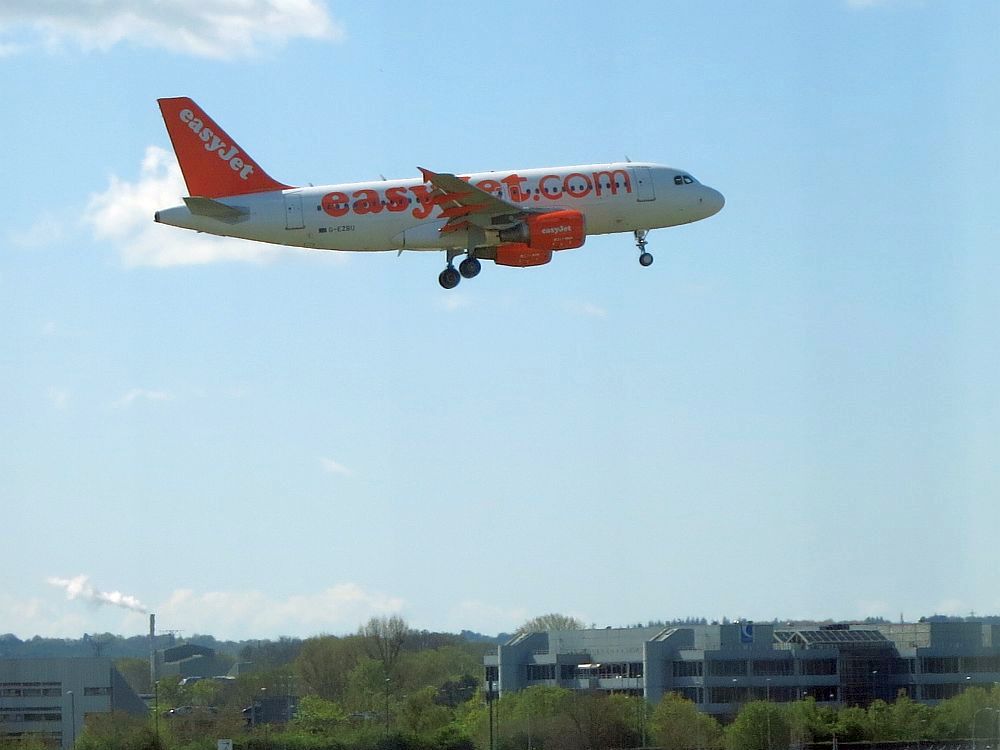 Against a blue sky, flat land at the bottom of the picture, wooded. Center top: an EasyJet plane comes into land, its landing gear almost fully lowered.