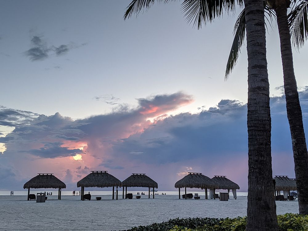 A view out to sea from the beach. On the right, two palm trees. white sand beach with a row of little thatched-roof shelters across the beach left to right. Beyond that, some dramatically-lit cloud formations over the ocean.