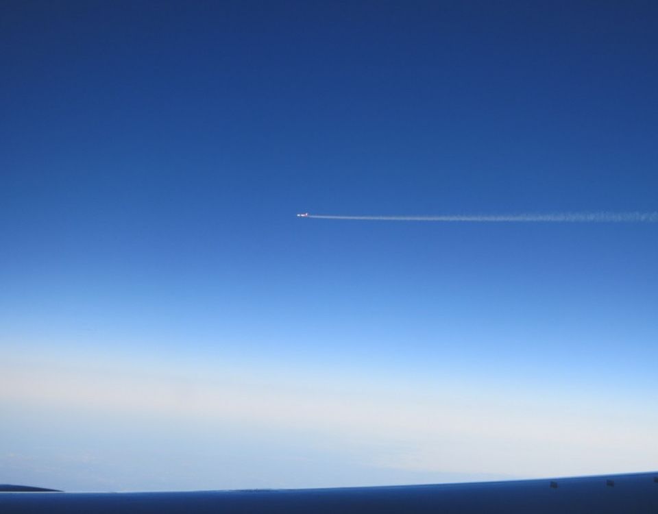 A deep blue sky, with the top of an airplain wing along the bottom of the photo. A tiny airplane in the center, with its smoke a horizontal line from the middle to the right edge of the photo