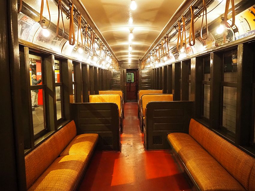 A view down the length of the inside of a train. The lighting is fairly low and yellowish. The seats are along the walls or perpendicular to it and are upholstered in a yellowish-orange fabric on the seats and backs. Straps hang in loops from the ceiling for standees to hang onto.