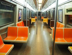 Looking down the length of a subway car. Harsh light from above, seats along the sides or perpendicular to the sides (in sets of 2). The seats are bright orange molded plastic.