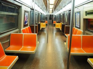 Looking down the length of a subway car. Harsh light from above, seats along the sides or perpendicular to the sides (in sets of 2). The seats are bright orange molded plastic.