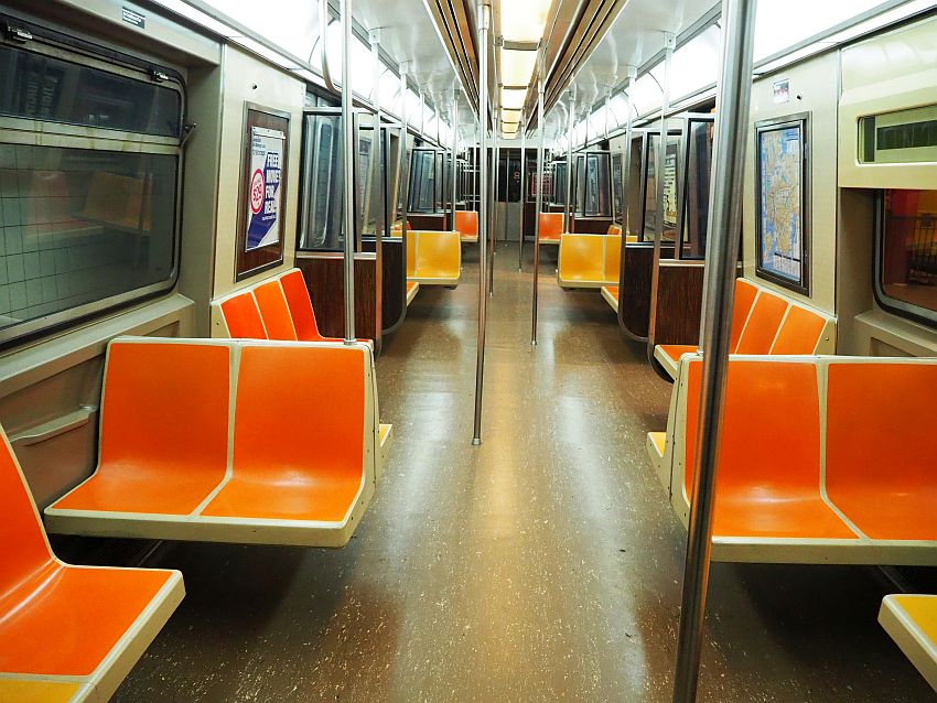 Looking down the length of a subway car. Harsh lighting, chairs of molded orange or yellow plastic along the walls or perpendicular to them. Here and there a floor to ceiling metal pole.