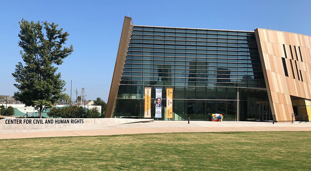 A modern building with a glass wall and wood-looking walls that lean inward.