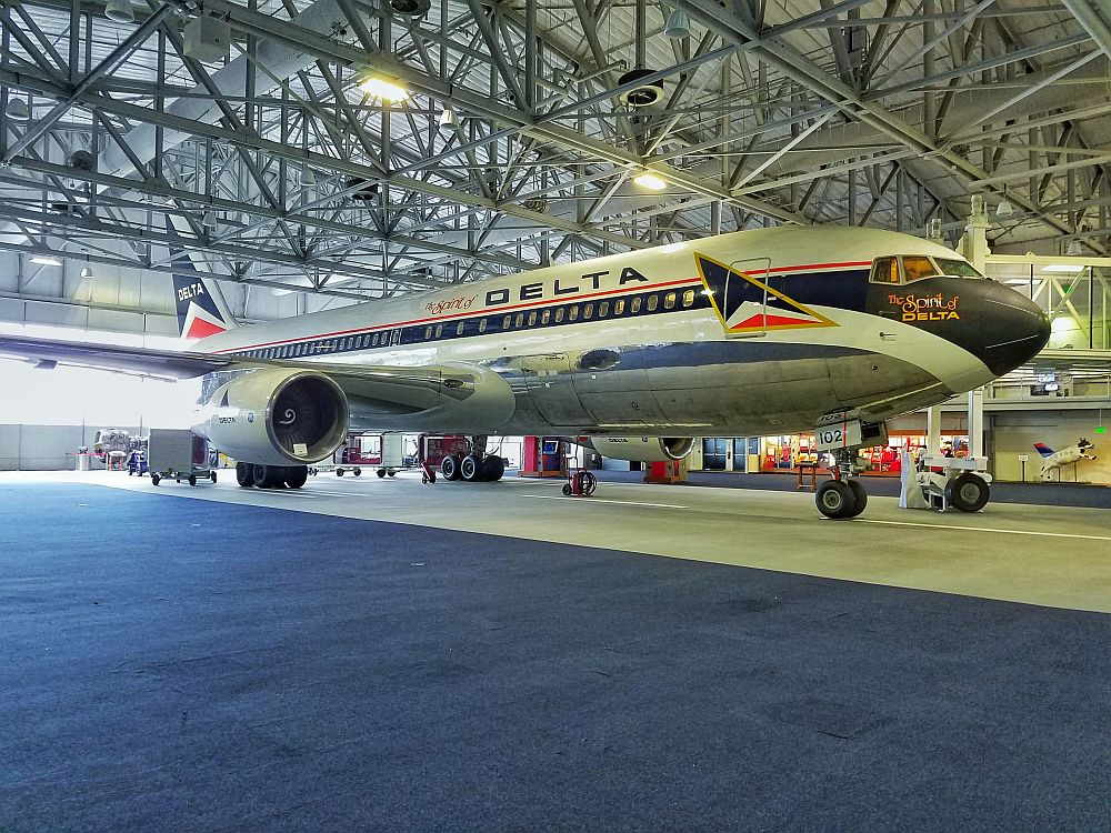 A large airplane inside a cavernous warehouse of a room (steel beams holding up a metal ceiling). The plane (a 737?) has the Delta logo and paintwork.