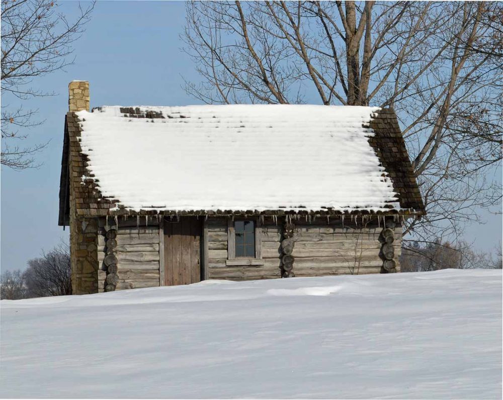 A simple log cabin with a slanted roof is one of the Little House on the Prairie locations. The roof has quite a bit of snow on it, with icicles hanging off the eaves and the field in front of the house is also covered in smooth snow. A large bare tree shows from behind the house.